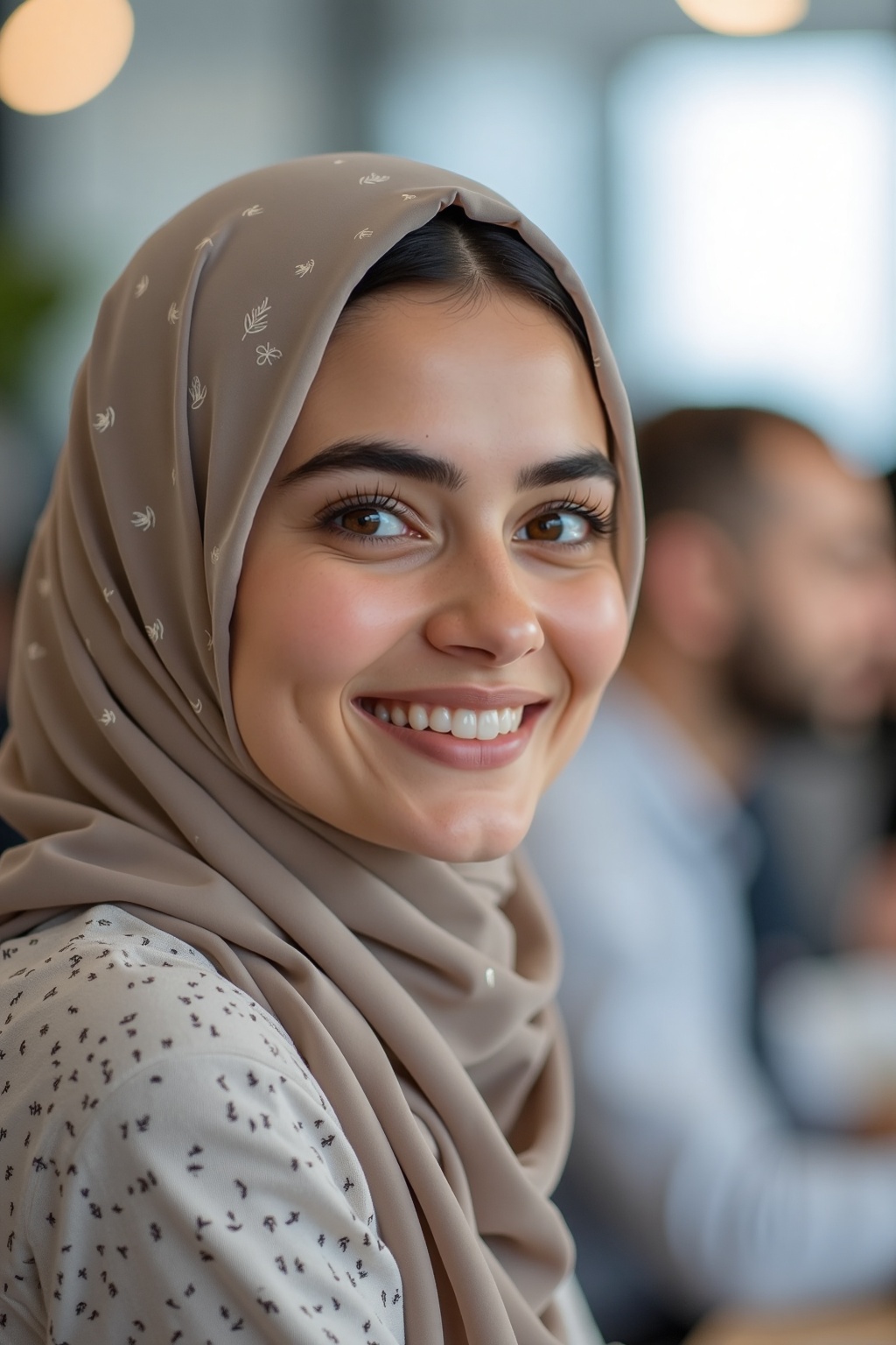In an office environment,  a young woman wearing a beige-patterned hijab casts a pleasant smile toward the viewer. Her pre...
