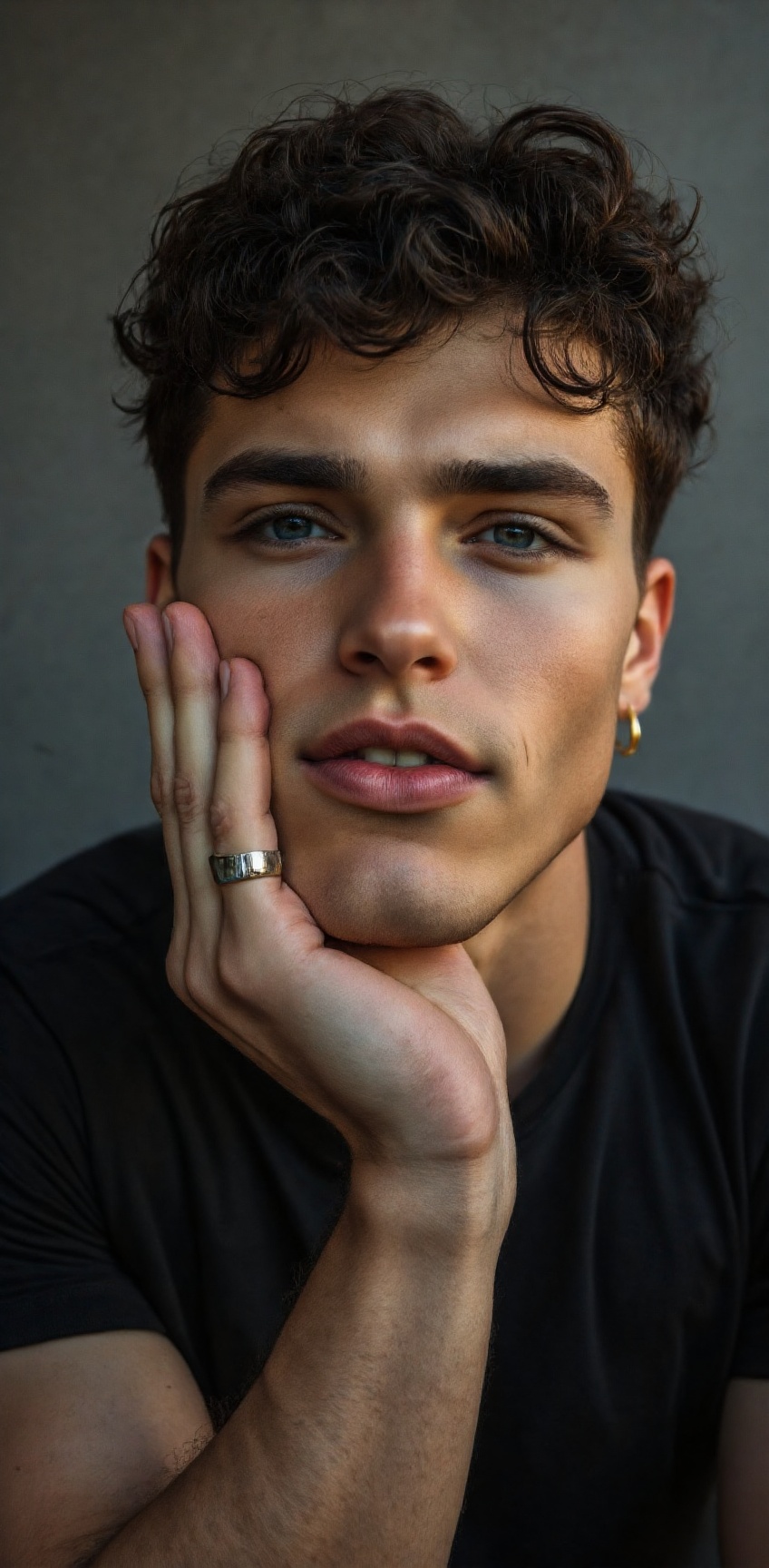 A close-up portrait of a young man with dark curly hair and blue eyes. He is wearing a black t-shirt and a silver ring on...