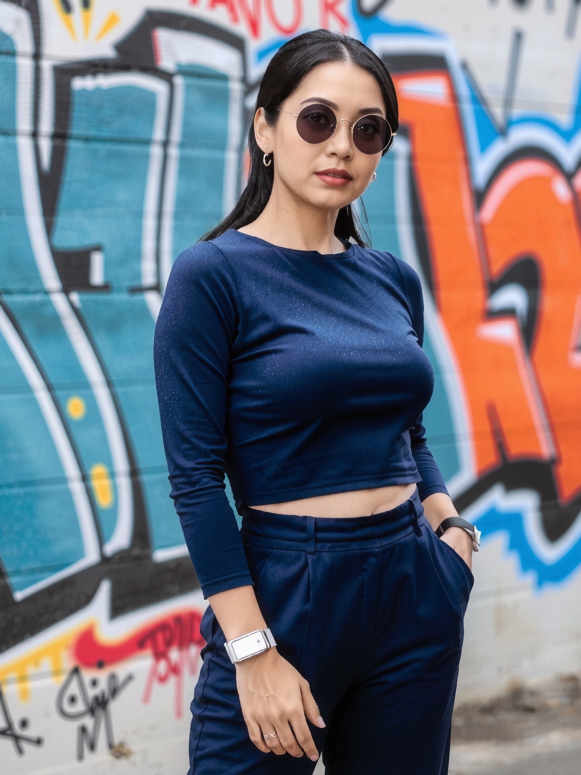 A woman stands confidently in front of a vibrant graffiti wall. She wears a chic,  navy blue ensemble featuring a midriff-...