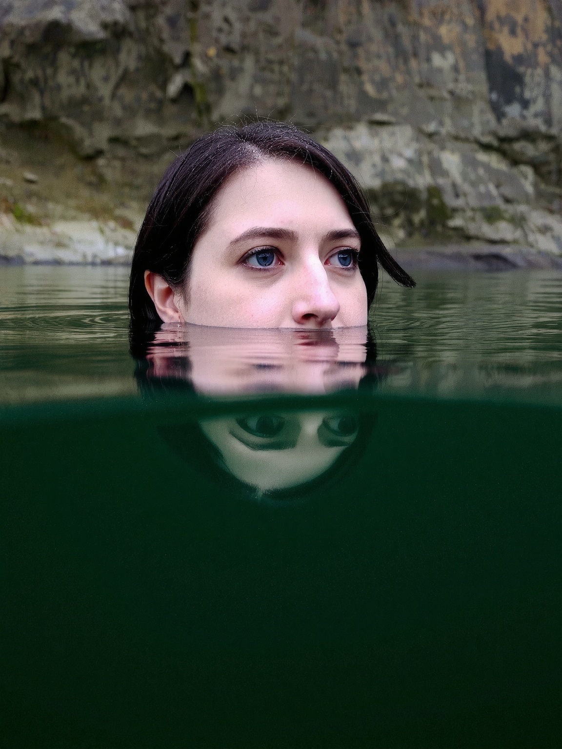 A woman is submerged in a body of water. The water is dark green. The woman's reflection is visible on the surface of the...