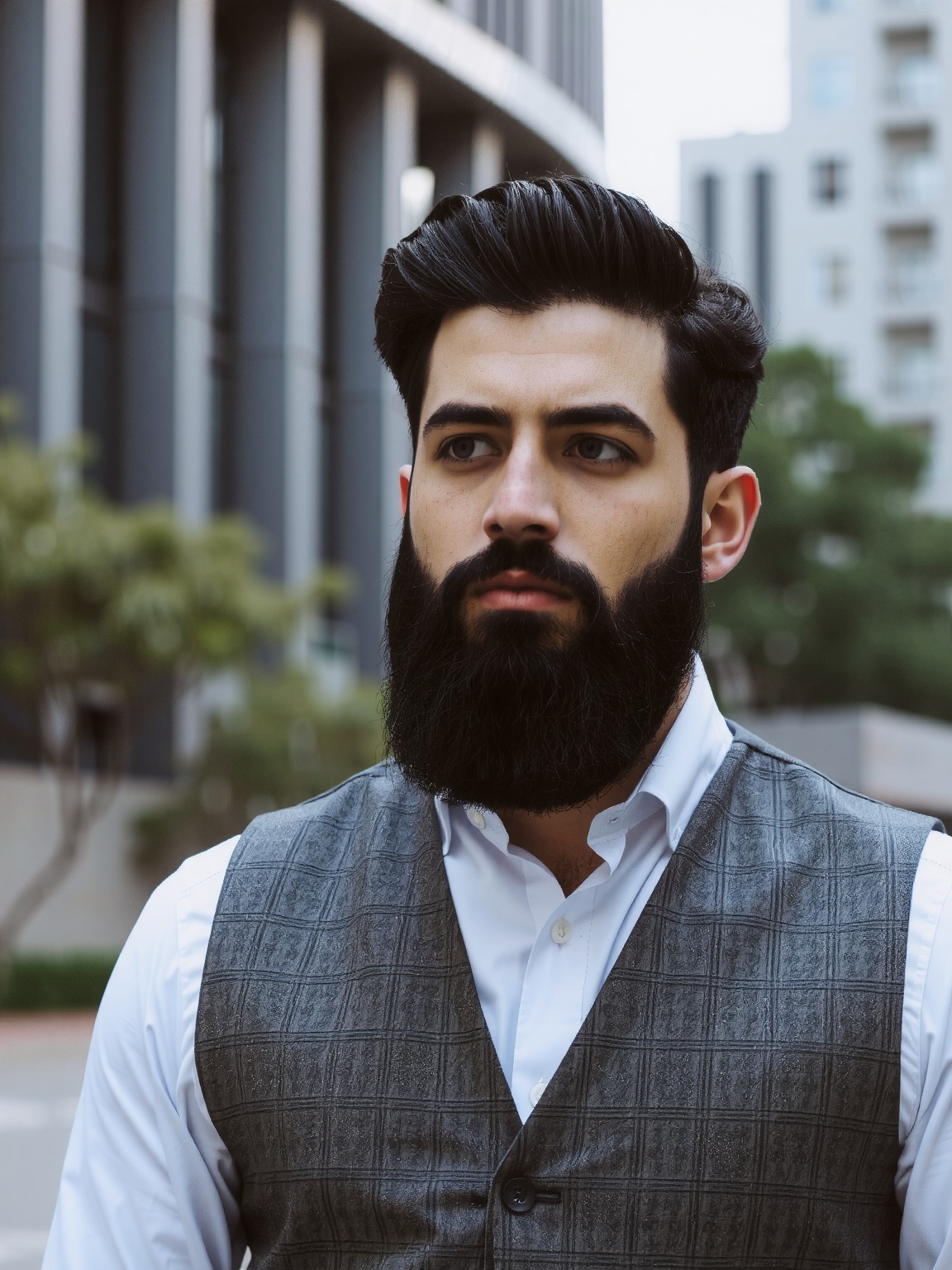 A man with dark,  neatly combed hair and a full beard stands against a modern building. He wears a white dress shirt and a...