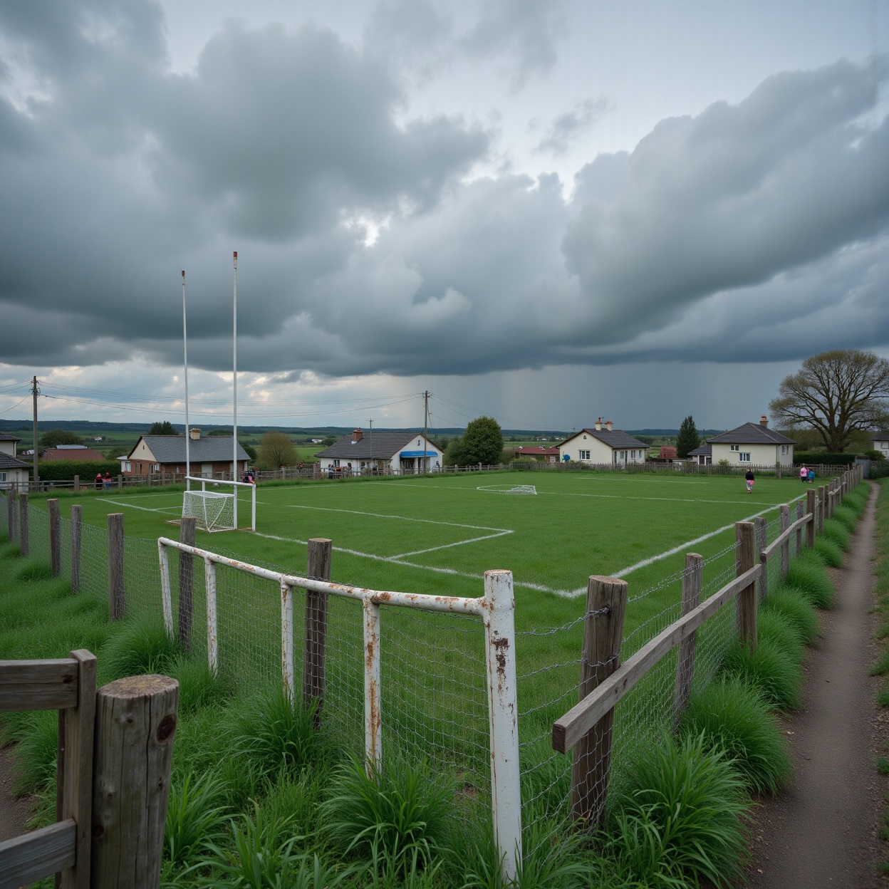 a rural football field under an overcast sky,  with dark storm clouds looming above. The field is surrounded by a simple w...