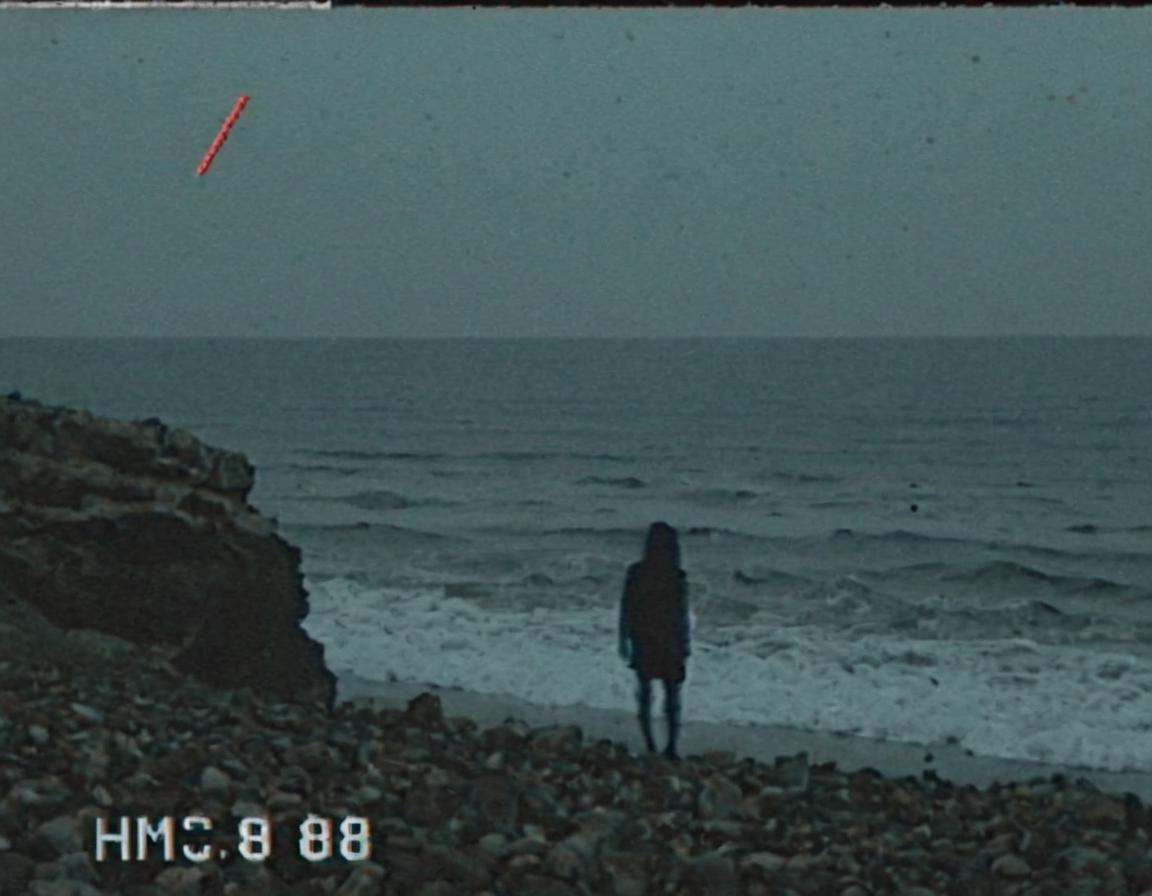 vhscore808,  VHS-style recording captures the silhouette of a lone figure standing at the edge of a rocky beach,  where oc...