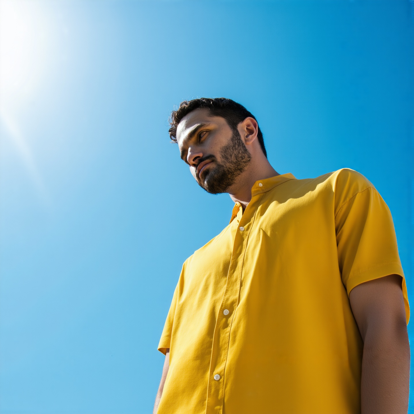 A man in a bright shirt stands against a clear blue sky. Soft sunlight highlights his relaxed posture. Clean lines and minimal design create a calm composition. Modern aesthetic.