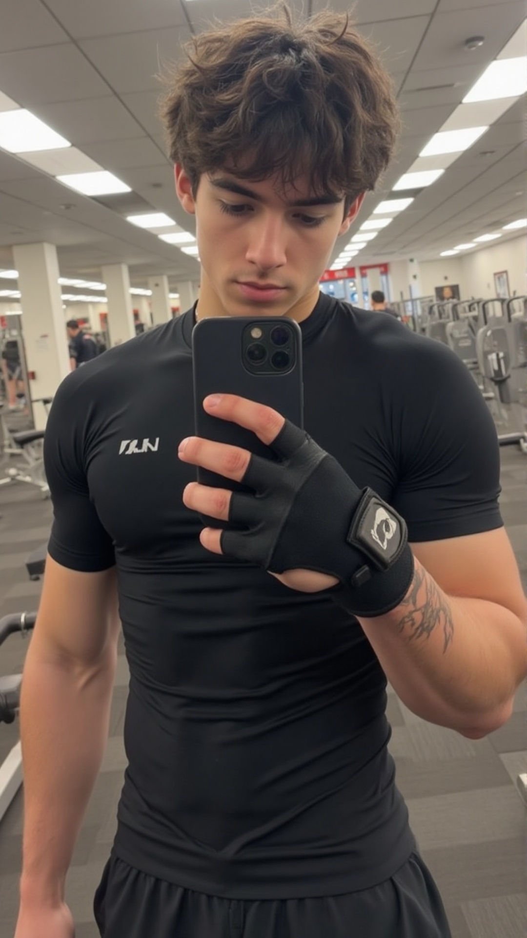 A young man with sharp features and tousled brown hair takes a gym mirror selfie,  his gaze slightly downward. He wears a...