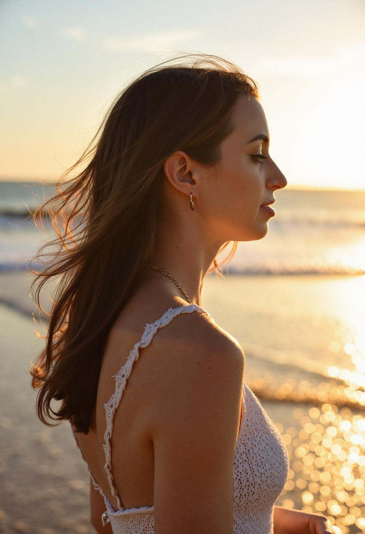 A woman in profile,  turned left,  standing on a beach at golden hour. Her hair gently moves in the breeze as she looks to...