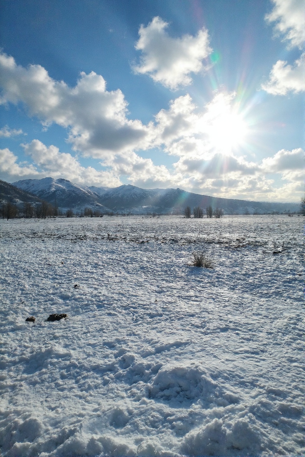 A panoramic view of a frozen moorland nestled between towering mountains. The sky is filled with distant clouds,  and sunl...