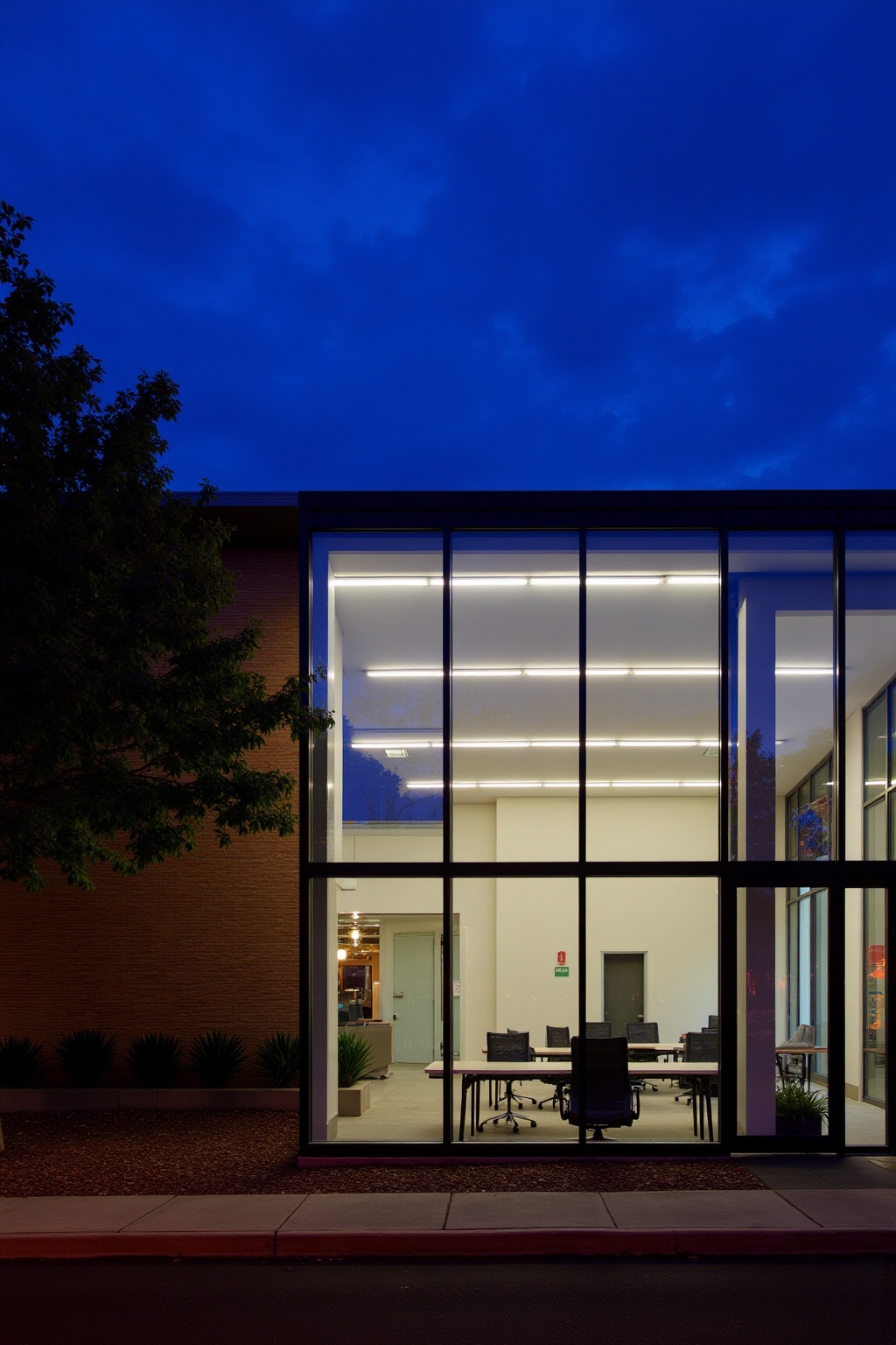 The image shows a modern building with large glass windows that reveal an interior workspace. The interior is brightly lit...