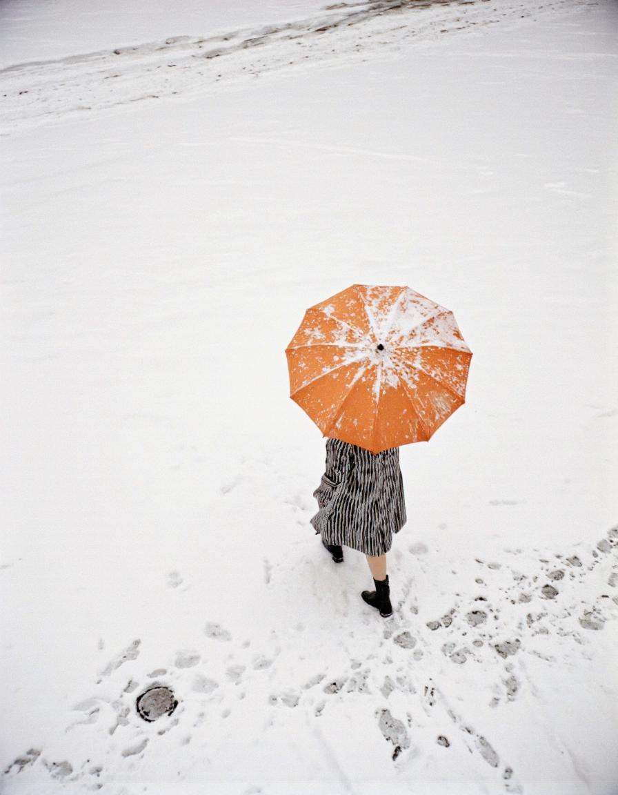 By Saul Leiter. A person walking alone in the snow,  holding an orange umbrella. The person is wearing a black and white s...