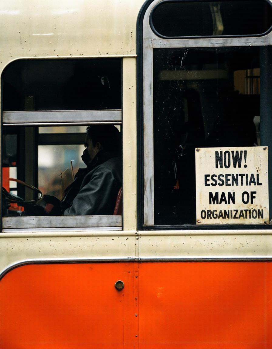 By Saul Leiter. A silhouette of a person sitting in the driver's seat of a bus,  viewed from behind. the bus is orange and...