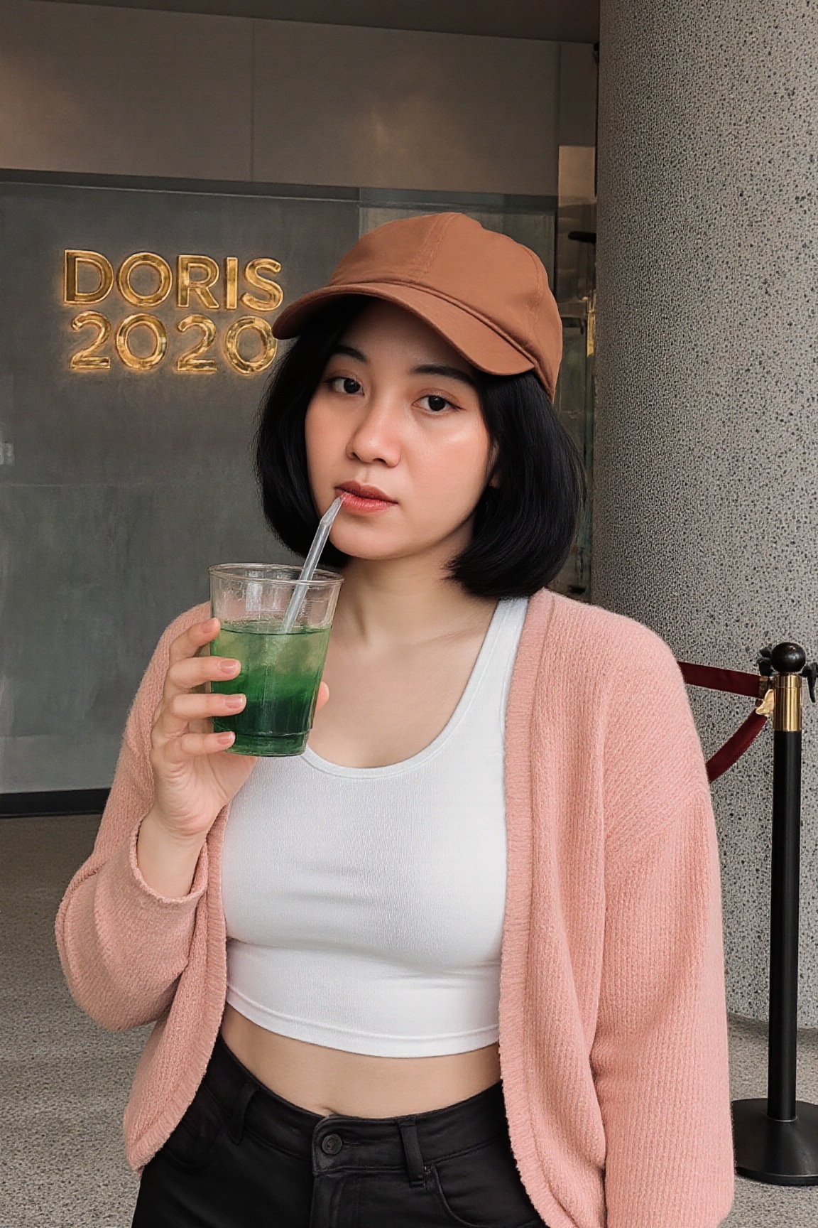 The image shows a woman standing indoors,  holding a green drink in a clear cup with a straw. She is wearing a white ****...