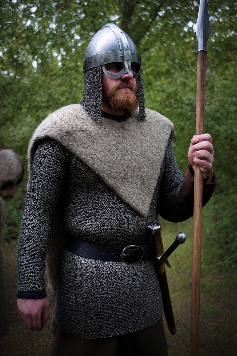 A photograph of a Viking warriors standing in a forest. The warrior has a reddish-brown beard and wears a steel spangenhel...